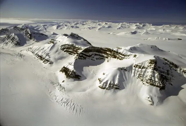 Pogled na snegom prekrivene planine i grebene koji se uzdižu iz ogromnog polarnog ledenog pokrivača, sa slojevitim stenovitim površinama vidljivim ispod belog snega i leda