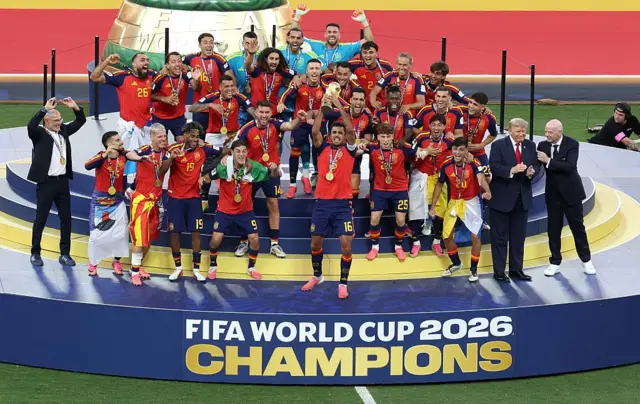كابتن المنتخب الإسباني، رودري، يرفع كأس العالم، فيما يظهر خلفه لاعبو المنتخب الإسباني وهم يرتدون الميداليات الذهبية ويحتفلون بالفوز. كما يظهر في الصورة الرئيس الأمريكي، دونالد ترامب، ورئيس الاتحاد الدولي لكرة القدم، جياني إنفانتينو، عقب فوز إسبانيا في المباراة النهائية لكأس العالم 2026.