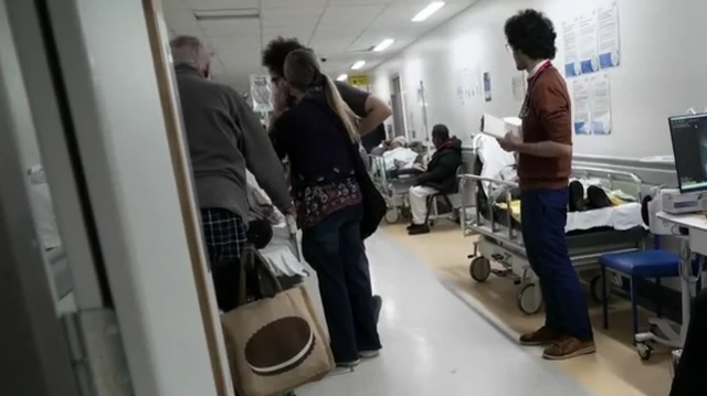 Los pacientes yacen en camillas a lo largo del pasillo de un hospital mientras la gente permanece de pie y camina entre ellos.