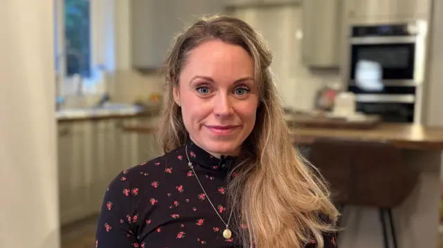 Niki Tibble, seated in a homely kitchen area, smiling at the camera. She is wearing a dark top with small red flowers on it. A gold-coloured locket-style necklace is around her neck. Her slightly wavy blonde hair is falling over her left shoulder and down her back. Behind her is a kitchen with a sink, cupboards and an oven, as well as an island unit with stools around it. 