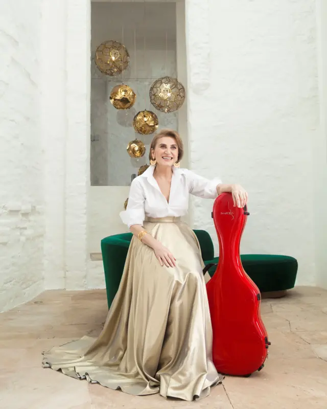 Berta Rojas posa en una sala. Está de pie y se apoya en un sofá verde y el estuche de su guitarra que es color rojo. 