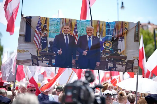 Zgromadzenie osób z biało - czerwonymi flagami. Na pierwszym planie transparent pokazujący stojących obok siebie Karola Nawrockiego i Donalda Trumpa