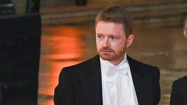 Morgan McSweeney, beyaz kravatlı bir kıyafet (siyah ceket, beyaz gömlek, kravat ve yelek) giymiş halde, arkasında yol ve sokak lambalarıyla görülüyor.