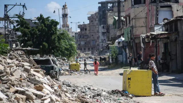 فلسطينيون يسيرون في شارع مليء بالأنقاض بجوار كتلة خرسانية صفراء تُشير إلى "الخط الأصفر" في 27 يوليو/تموز 2026 في حي التفاح بمدينة غزة. 