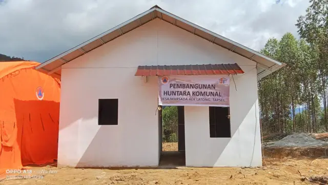Satu unit huntara yang cukup besar, berdinding multiplek dan rangka besi, berdiri di sebelah tenda oranye BNPB. Di depan rumahnya ada spanduk bertuliskan, "Pembangunan huntara komunal Desa Marsada Aek Latong, Tapsel".