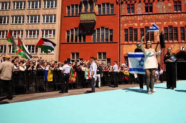 Basel'de açık havada yapılan tanıtım etkinliğinde Filistin bayrağı açan kişiler, kurulan podyumda yürüyen ve elinde İsrail bayrağı taşıyan şarkıcıyı protesto ediyor.