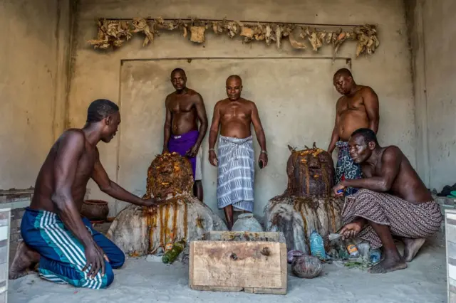 Des hommes, torses nues, autour de deux monticules de terres sur lesquelles sont versée de l'huile rouge.