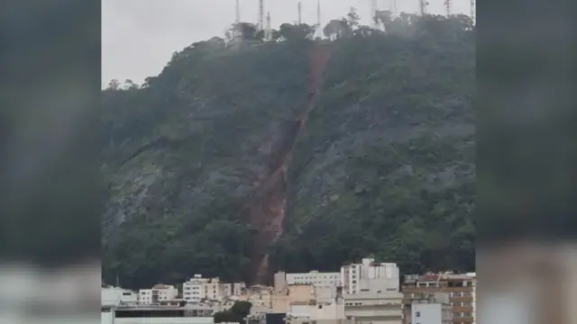 岩や植生で覆われた鋭い傾斜の斜面に、茶色い土がほぼ一直線に流れ落ちた様子が見える。その手前下には白いビルなどが並んでいる