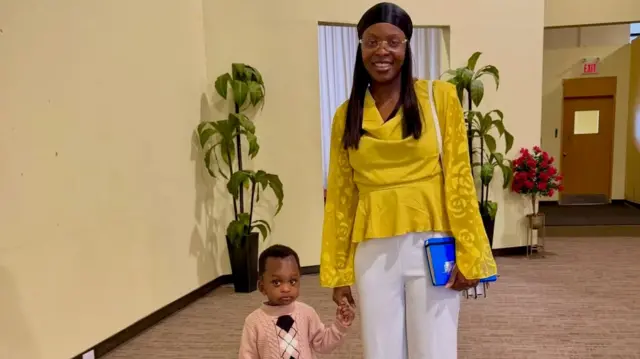 Ngozi Okobi-Okeoghene porte un chemisier jaune moutarde, un pantalon blanc, un foulard noir et des lunettes. Elle tient un livre bleu à la hanche gauche. De la main droite, elle tient la main d'un petit garçon vêtu d'un pull rose pâle. Tous deux regardent l'objectif. La scène se déroule dans un hall d'entrée spacieux, recouvert de moquette, aux murs aux tons neutres, agrémenté de deux plantes vertes en pot, d'une composition florale rouge et, en arrière-plan, de ce qui semble être une porte coupe-feu.