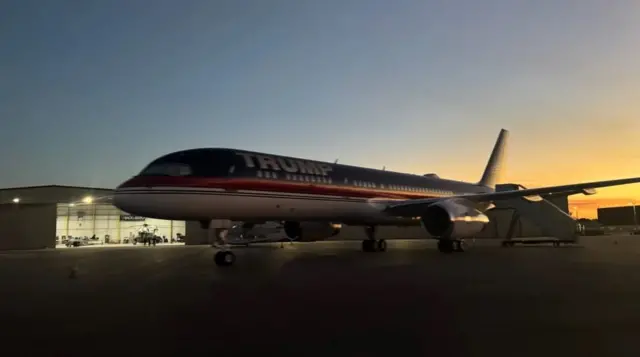 Пасажирський літак, припаркований на аеродромі біля ангара. На борту літака великими літерами написано "TRUMP". Літак стоїть на тлі вечірнього або ранкового неба з теплими відтінками. На передньому плані видно шасі літака, а поруч – трап для посадки. У глибині кадру – ангар і ще один невеликий літак і гелікоптер всередині.