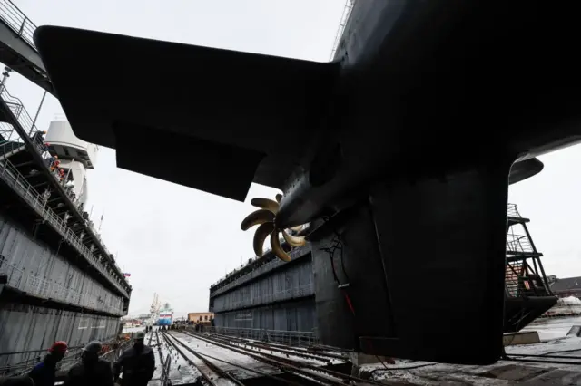 Великий підводний човен у сухому доку, видно його кормову частину з великим гвинтом і вертикальним рулем. Навколо – металеві стінки доку, рейки для переміщення судна, кілька людей у захисних касках. Освітлення природне, небо похмуре.