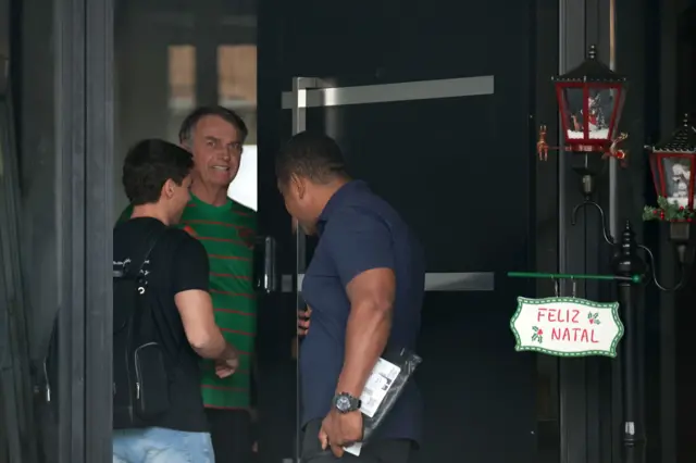 Bolsonaro sorrindo ao abrir a porta de casa para dois homens