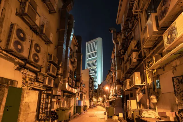 Sebuah jalan sempit dengan banyak AC dan gedung pencakar langit modern di latar belakang pada malam hari.