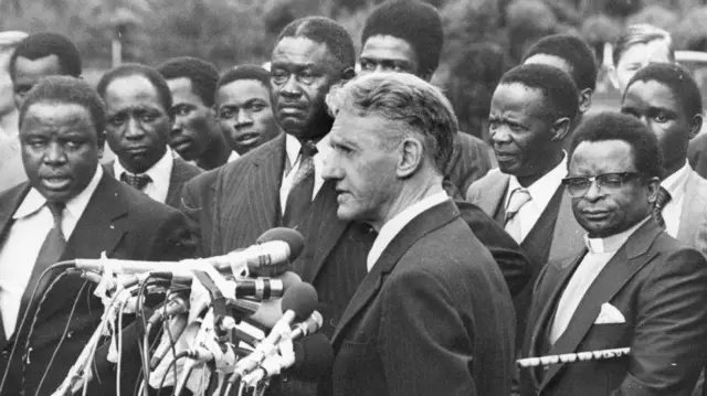 Firaministan Rhodesia Ian Smith tsaye a gaban makirofon bayan wani taro da Dakta David Owen sakataren harkokin wajen Birtaniya da Cyrus Vance sakataren harkokin wajen Amurka.