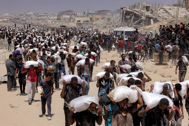 فلسطينيون يسيرون حاملين أكياس دقيق تم تسليمها بعد دخول شاحنات محملة بالمساعدات الإنسانية إلى شمال غزة في 27 يوليو/تموز 2025 قادمة من معبر زيكيم الحدودي.