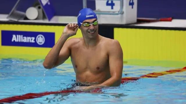 A foto mostra um nadador em uma piscina com uma touca azul e óculos de natação. 