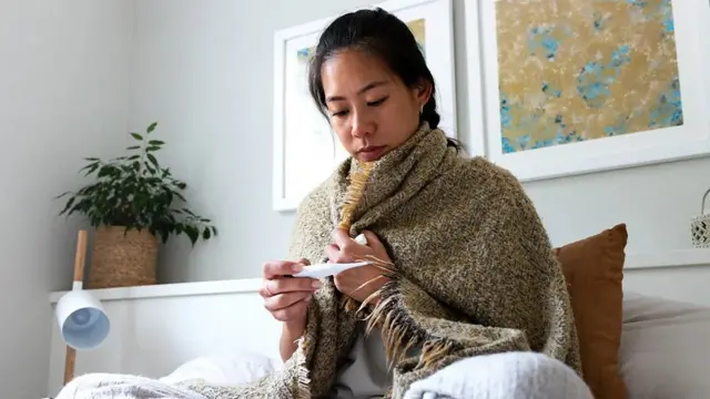Une femme assise emmitouflée sous une couverture en laine marron pose un thermomètre blanc contre sa peau