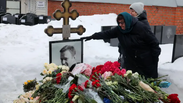 Людмила Навальная прикасается к фотографии своего сына на нагробии, на могиле лежит много букетов цветов