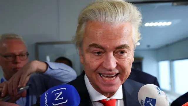 白髪をオールバックにしたスーツ姿の男性が、記者に囲まれながら話している。男性の前には青や白、黒のマイクが掲げられている。後ろからカメラやスマートフォンを構える人もいる