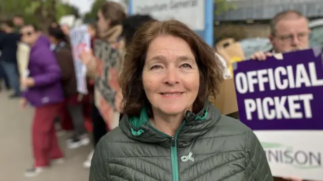 A woman with brown mid-length wavy hair smiles at the camera in front of a picket line. She is wearing a green puffa jacket with turquoise zip. The people behind her are holding placards and wearing burgandy scrubs.