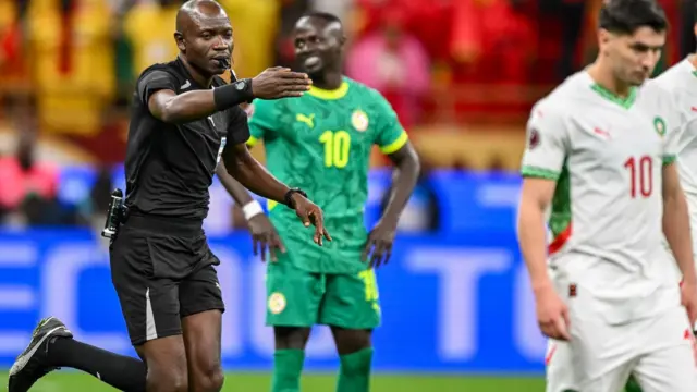 L’arbitre Jean‑Jacques Ndala, vêtu d’une tenue entièrement noire et le sifflet entre les lèvres, désigne le point de penalty de la main droite tout en courant derrière Brahim Diaz du Maroc, habillé en blanc avec des touches rouge et verte, après avoir accordé un penalty controversé au Maroc lors de la finale de la CAN 2025. En arrière‑plan, Sadio Mané du Sénégal, en tenue entièrement verte, se tient les mains sur les hanches et regarde vers sa droite avec un sourire en coin.