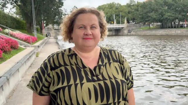 میرکا موکونن در کنار رودخانه هاله ایستاده و مستقیماً به کمره نگاه می‌کند و لباسی طرح دار سبز و مشکی پوشیده است. او موهای مجعد بلوند کوتاهی دارد