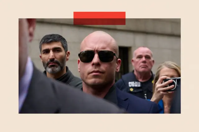 A bald white man in sunglasses and a navy blue suit frowns. He is surrounded by people, including a police officer.