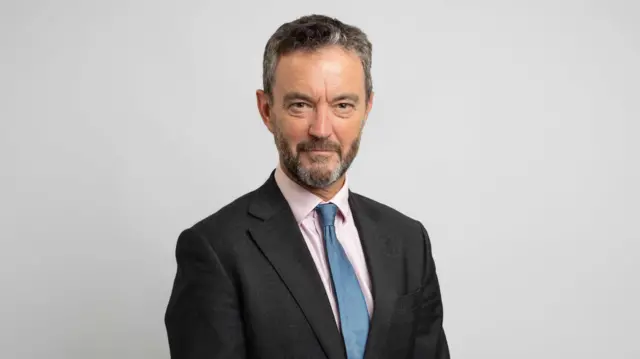 Matthew Jowitt sonríe y mira a la cámara frente a un fondo gris. Tiene el pelo corto y canoso, barba y viste una camisa negra sobre una camisa rosa con corbata azul.