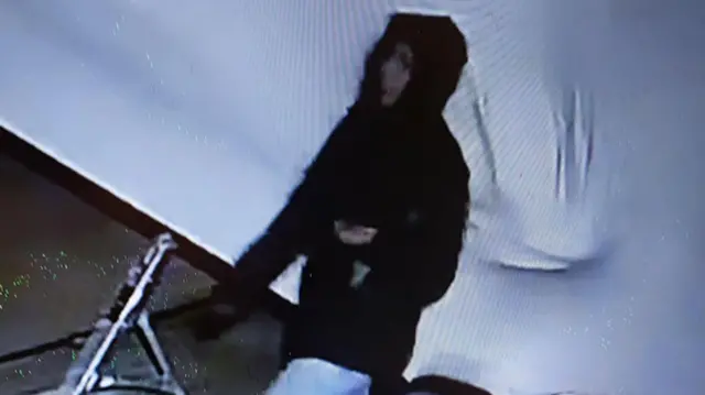 Imagem de uma câmera de segurança mostra uma pessoa com um casaco preto com capuz caminhando.