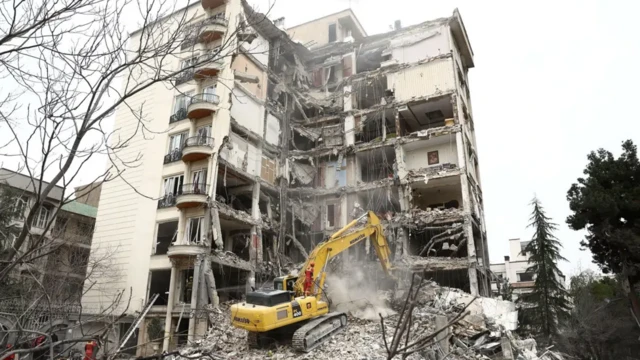 یک ساختمان هشت طبقه نیمه ویران شده و یک بیل مکانیکی در کار آواربرداری