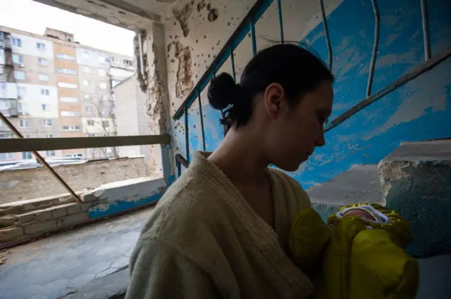 В під’їзді з пошкодженими стінами стоїть жінка у домашньому халаті, яка тримає на руках немовля в теплому жовтому одязі. Стіни та сходи вкриті слідами уламків і облущеною фарбою. Крізь велику діру у зовнішній стіні видно двір і багатоповерхівку навпроти.