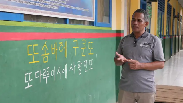 Abidin, guru yang mengajar aksara Hangeul di Baubau, berdiri di sebelah kalimat bertulis, "Kita hormati guru kita, sayangi guru kita" dalam bahasa Cia-Cia, tapi ditulis dalam aksara Hangeul.