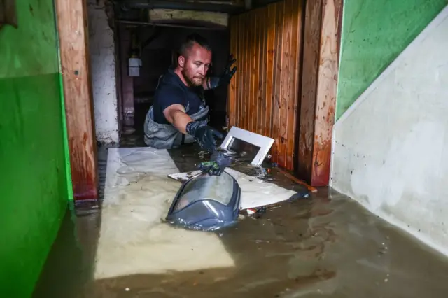 Чоловік стоїть у дверях по груди у воді, викидаючи назовні речі, які плавають серед брудної води.
