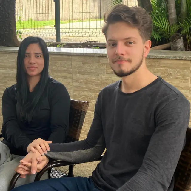 Casal sentado, com as mãos dadas, diante de mesa em área externa