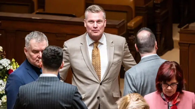 Чоловік у бежевому костюмі та краватці дивиться в камеру, поряд з ним кілька людей