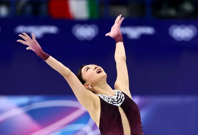 首元に白いパールがついた、ボルドーのホルターネックドレス風衣装を着た坂本が、両手を広げ、笑顔で上を見ている