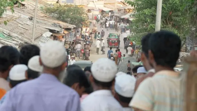 কক্সবাজারে রোহিঙ্গা ক্যাম্পের একটি রাস্তার ছবি, টুপি পরা কিছু মানুষ জড়ো হয়ে তাকিয়ে আছেন রাস্তার দিকে