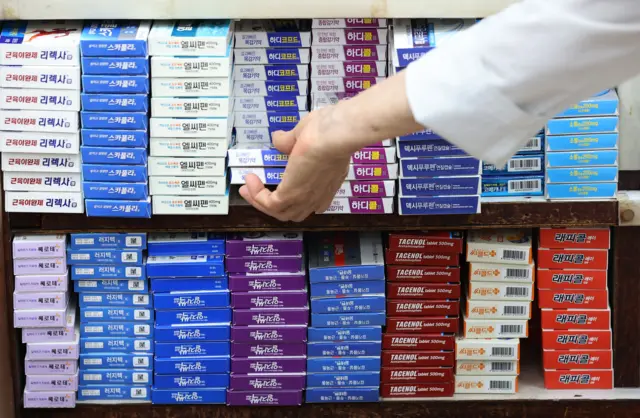 약국 진열대에 각종 감기약이 진열되어 있다. 약사의 손이 그 중 하나를 집어 들고 있다