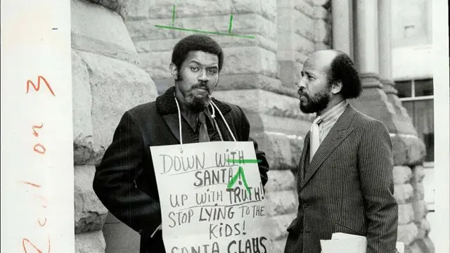 「サンタを倒せ！」「子どもにうそをつくのをやめろ！」などとかかれたサインを首から下げた、ヴェトナム戦争の退役軍人リチャード・ディルディ氏の白黒写真。黒人のディルディ氏はフォーマルなシャツにネクタイを着けている。左隣にはディルディ氏を見つめる男性が立っている