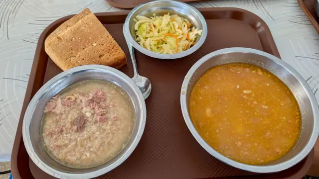 Три металлические миски на подносе с куском хлеба. В двух — супы, в одной с мясом, а в третьей — салат.