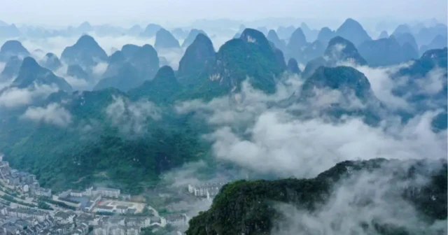 A imagem mostra uma paisagem natural composta por montanhas cobertas por vegetação, com cumes pontiagudos que emergemcasas de apostascasas de apostas em dolardolaruma névoa espessa, criando uma atmosfera etérea. Na parte inferior, há um pequeno aglomerado urbano com prédios e ruas, que contrasta com a imensidão das formações naturais. O céu tem tons azulados, reforçando o climacasas de apostascasas de apostas em dolardolarserenidade e mistério da cena.