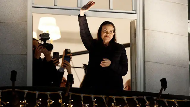 Siyah giymiş bir kadın otel balkonundan el sallıyor, arkada kaldığı odada fotoğrafçılar var