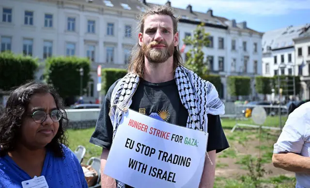 متظاهر أوروبي يحمل لافتة كتب عليها "المجاعة تضرب غزة. أيها الاتحاد الأوروبي! أوقف التجارة مع إسرائيل"، وذلك في وقفة احتجاجية في أمام البرلمان الأوروبي في يونيو/حزيران ٢٠٢٥ في بروكسل، بلجيكا.