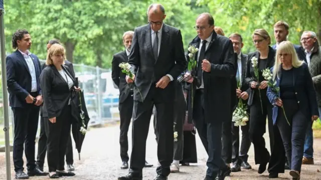 Almanya Başbakanı Merz elinde çiçek demeti ve koyu renk takım elbiseli diğer görevlilerle olay yerini ziyaret etti