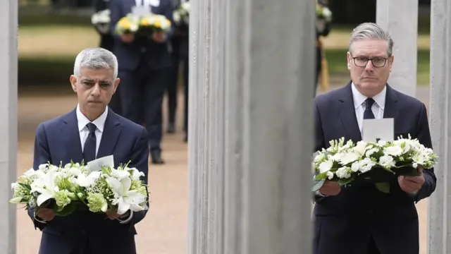 紺色のスーツに白いシャツを着たグレーのショートヘアのサディク・カーン・ロンドン市長と、眼鏡をかけ黒いスーツを着たグレーの髪のキア・スターマー首相が、白い花輪を持って灰色の石柱の両側を歩いている