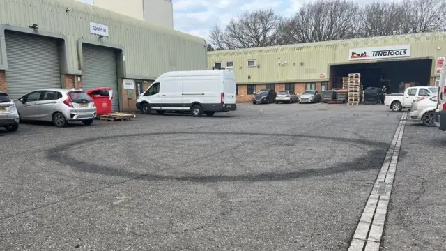 Una carretera y un aparcamiento en un polígono industrial con un edificio de dos plantas al fondo y un edificio de una sola planta con grandes persianas a la izquierda. Hay coches y furgonetas aparcados cerca de los edificios. Un gran círculo negro (una marca de derrape) se encuentra en el centro de la imagen. Se ven más círculos tenues a su alrededor.