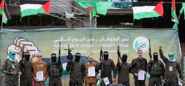 Foto dos reféns ao lado de soldados do Hamas em um palco. 