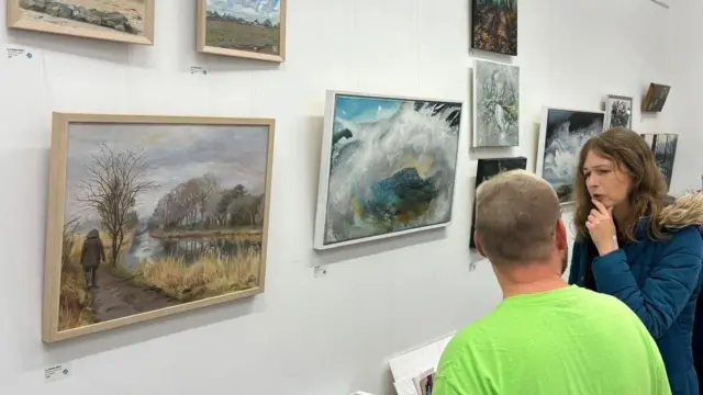 Dentro de una galería de arte con numerosas pinturas en la pared, un hombre con una camiseta verde claro conversa con una mujer de cabello castaño largo que lleva un abrigo azul con capucha de piel.