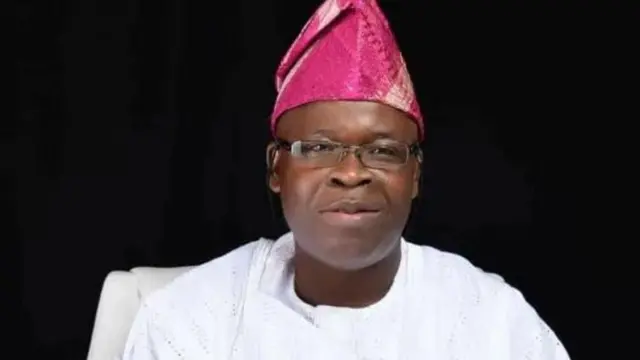 Aworan Oloye Dotun Sanusi ree. O wọ agbada funfun, o de fila alawọ pupa o si fi igo soju. 