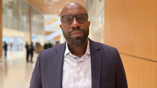 Maître Richard Gisagara uburanira abareze Charles Onana. Ahagaze hanze y'urukiko i Paris mu Bufaransa, yambaye indorerwamo, ishati ijya kuba umweru n'ikoti rijya kuba umukara, inyuma ye hahagaze abantu batagaragara neza. Ifoto yo ku itariki ya 9/12/2024